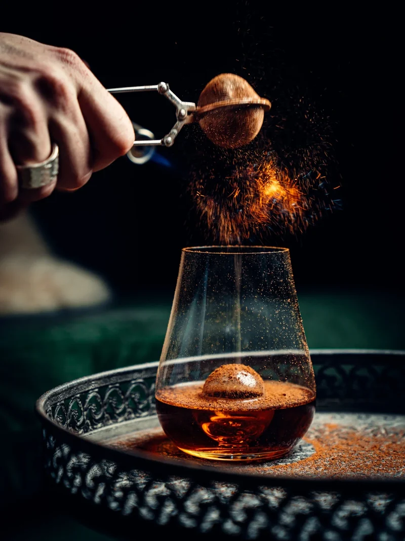 Hand streut Gewürz über einen Cocktail vor dunklem Hintergrund