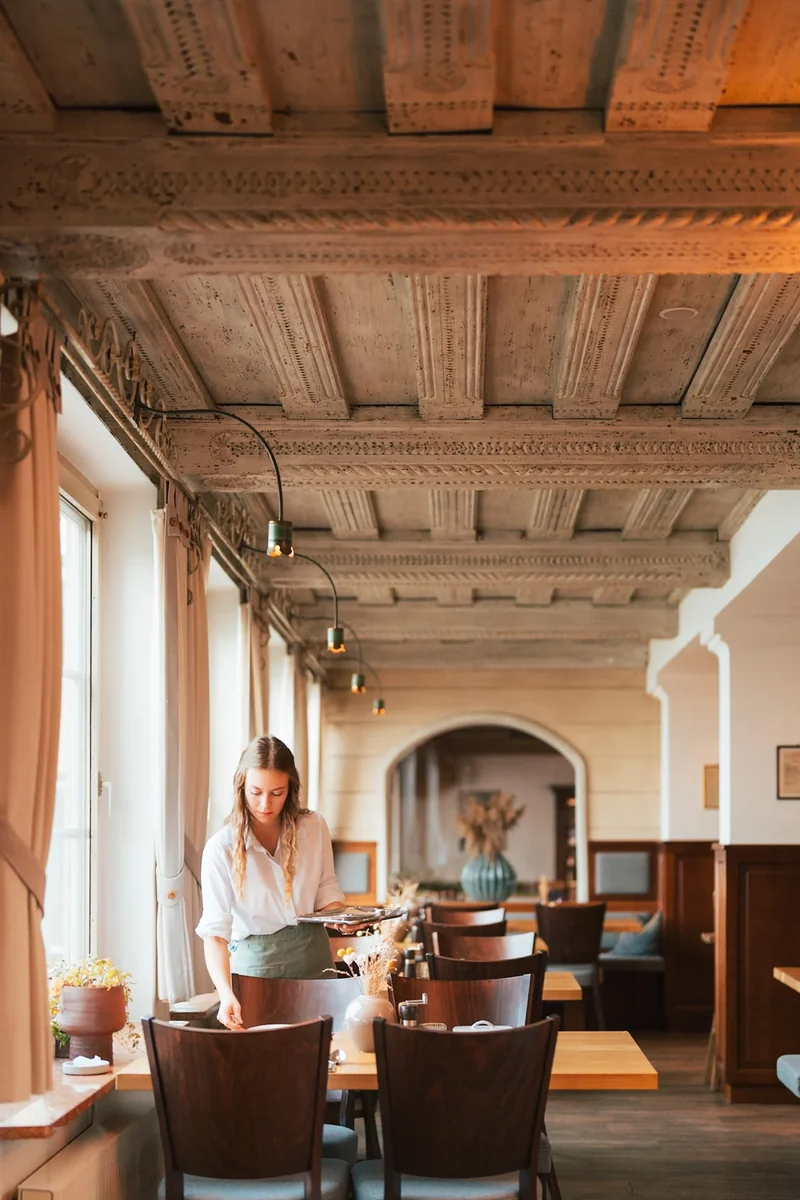 Mitarbeiterin im Restaurant mit Holzbalken und warmem Fensterlicht